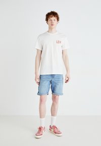 Λευκό βαμβακερό μπλουζάκι με το λογότυπο "LEE" σε κοραλί, συνδυασμένο με ανοιχτό μπλε τζιν σορτς με διπλωμένα μανίκια, λευκές κάλτσες και ροζ αθλητικά παπούτσια.