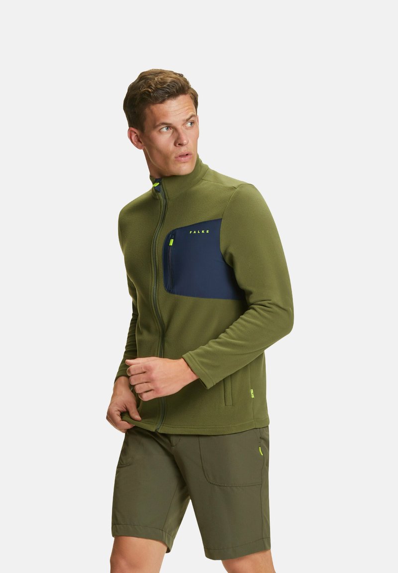 Giacca in pile verde con tasca anteriore blu navy, chiusura a zip e colletto alto; abbinata a pantaloni corti color oliva, entrambi con accenti gialli vivaci.
