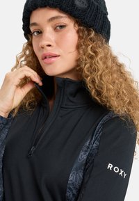 Frau mit lockigem Haar, die eine schwarze Strickmütze und eine schwarze Jacke mit Reißverschluss und gemustertenÄrmeln trägt, auf denen "ROXY" steht, während sie den Kragen anpasst.