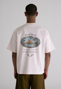 Light pink cotton t-shirt featuring a panoramic resort illustration on the back, surrounded by ornate black detailing and text.