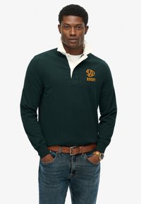 Camiseta de rugby de manga larga en verde oscuro con un cuello blanco y texto bordado en amarillo que dice "RUGBY". Se lleva con un cinturón marrón y jeans azules.