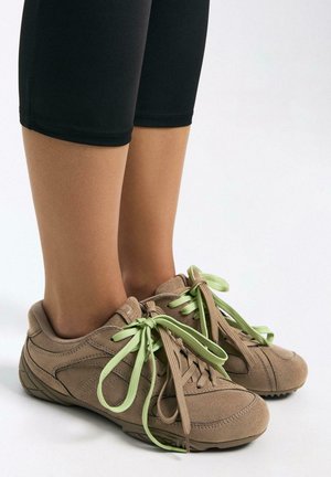 Nohy v černých legínách a béžových semišových teniskách s kombinovanými béžovými a světle zelenými tkaničkami, stojící na bílém povrchu.