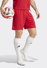 Rote Sportshorts mit weißen seitlichen Akzenten, kombiniert mit knielangen weißen Socken mit schwarzem Logo und weißen Fußballschuhen, während er einen Fußball hält.