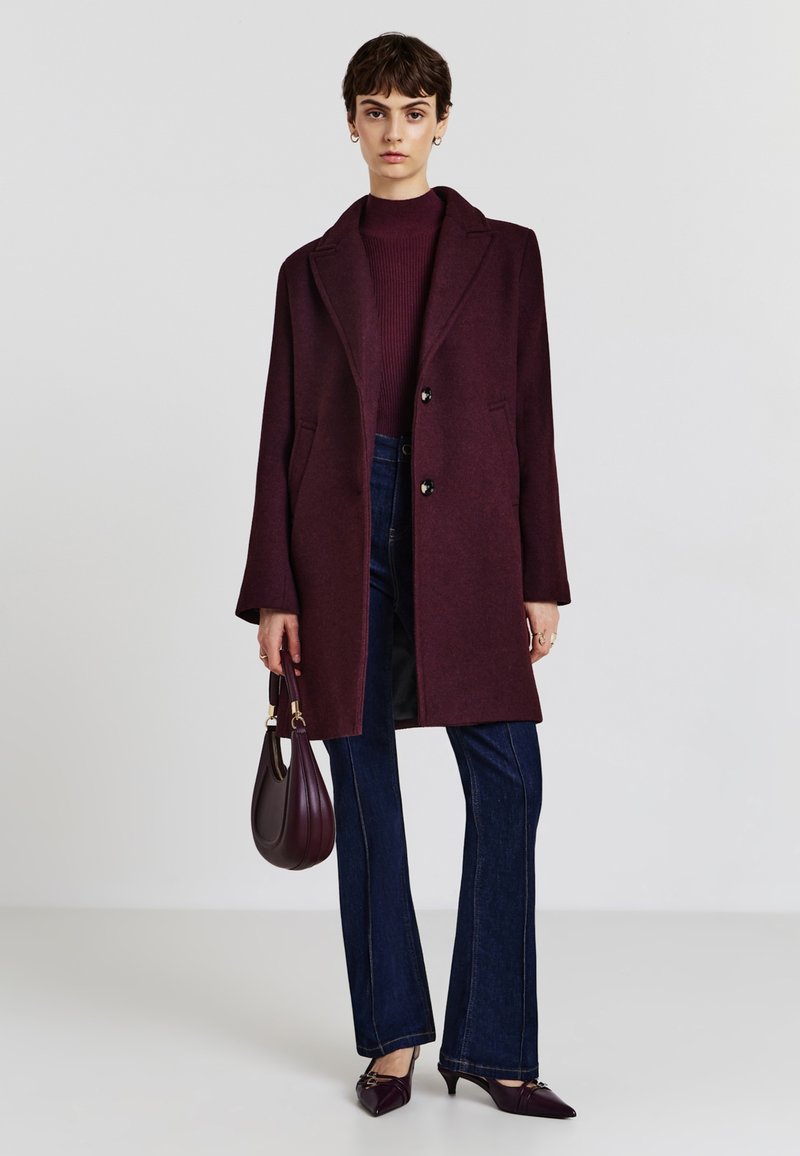 Burgundy wool coat with notch lapels, two buttons, and side pockets; paired with dark blue flared jeans and pointed-toe heels.