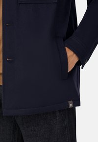 Marinblå tygjacka med texturerad yta, sidoficka och knäppning med knappar. Har en liten logotags på den nedre kanten.