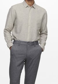 Uomo che indossa una camicia a maniche lunghe beige chiaro, infilata nei pantaloni grigi su misura, con le mani nelle tasche, su sfondo bianco.