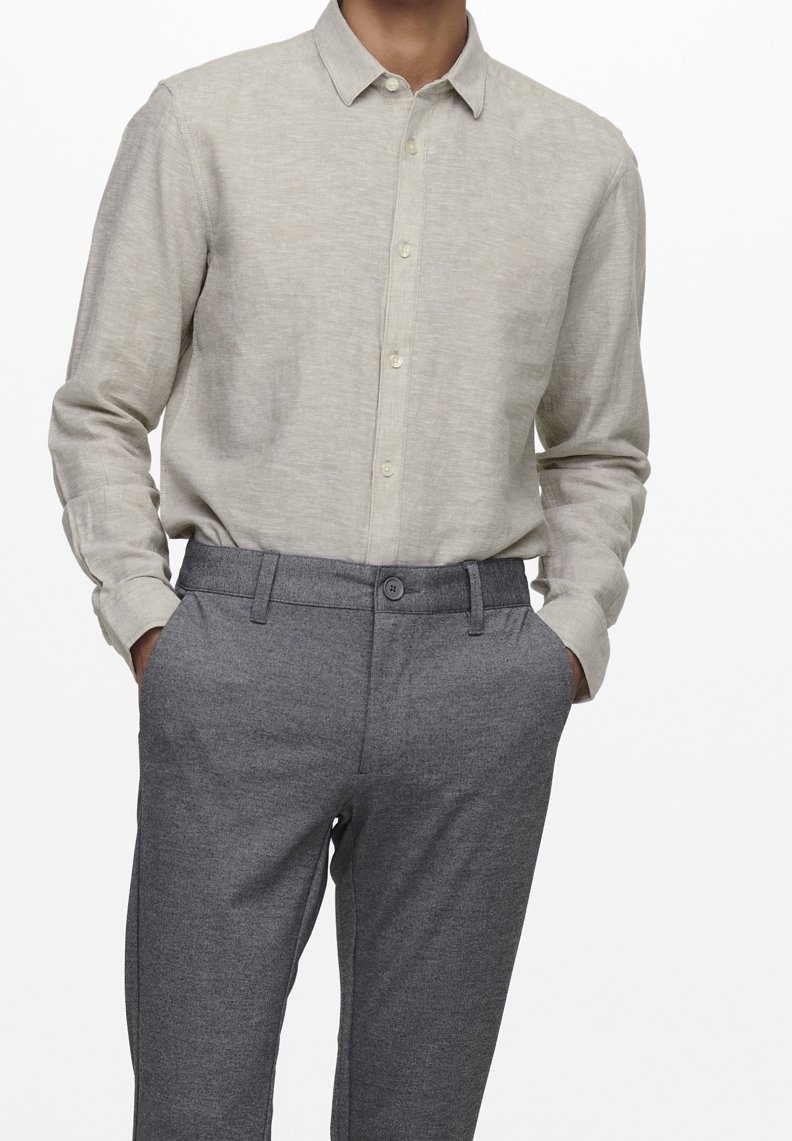 Uomo che indossa una camicia a maniche lunghe beige chiaro, infilata nei pantaloni grigi su misura, con le mani nelle tasche, su sfondo bianco.