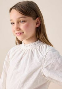 Jeune fille aux cheveux châtain clair portant une blouse blanche brodée, souriant doucement, sur un fond beige uni.