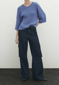 Personne portant un pull en tricot bleu ample et un pantalon cargo en denim foncé à jambes larges, debout devant un fond uni.