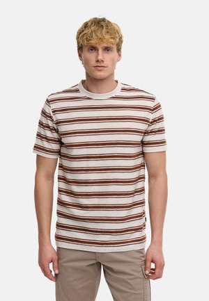 Blonder Mann, der ein beige und braun horizontal gestreiftes kurzärmeliges Hemd und hellbraune Hose trägt, steht vor einem schlichten Hintergrund.