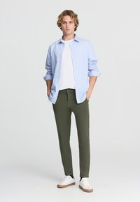 Jasnoniebieska koszula na guziki noszona na białym t-shircie, w połączeniu z oliwkowo-zielonymi spodniami slim-fit i białymi trampkami z brązowymi akcentami.