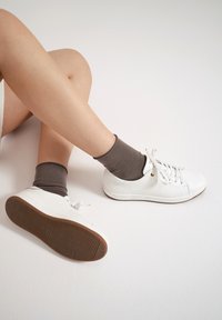 Białe skórzane trampki z okrągłym noskiem, teksturowaną powierzchnią, płaską brązową podeszwą i sznurowanym designem. Połączone z ciemnobrązowymi skarpetami.