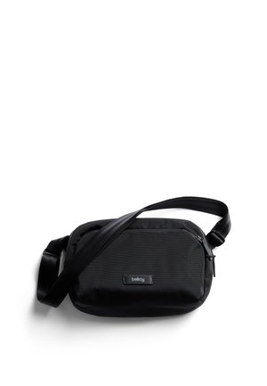 Sac bandoulière noir en nylon à texture lisse, doté d'un patch de marque et d'une sangle réglable. Forme rectangulaire compacte.