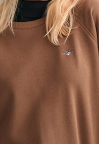 Brązowy bawełniany sweatshirt z okrągłym dekoltem i ściągaczami przy mankietach. Posiada małe haftowane logo na piersi. Gładka tekstura.