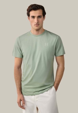 Junger Mann trägt ein hellgrünes kurzärmeliges T-Shirt mit kleinem Logo auf der Brust und weiße Hosen, steht vor einem neutralen Hintergrund.