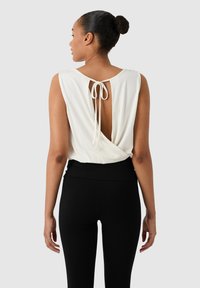 Femme portant un chignon, vêtue d'un haut sans manches blanc avec un détail en trou de serrure au dos et un lien, associé à un pantalon noir taille haute.