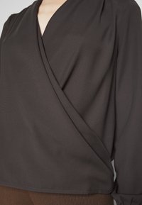 Blouse portefeuille marron avec col en V, manches longues, texture douce et fronces aux épaules.