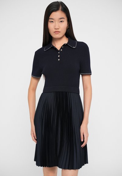 Laivastonsininen polo-mekko, jossa on ribbitetty yläosa ja laskostettu hame. Ominaisuuksiin kuuluu kaulus, viiden napin läppä ja kontrastiväriset yksityiskohdat. Tasainen rakenne kauttaaltansa.