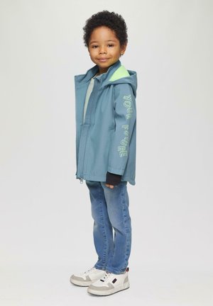 Blue jacket with a hood, featuring green accents and “YOUR FUTURE” text on the sleeve. Worn over a light shirt and blue jeans. White sneakers.