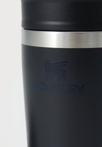 Čierna nehrdzavejúca oceľová cestovná hrnček s hladkým matným povrchom, strieborný prstenec blízko vrchu a embosované logo "STANLEY" s dizajnom leva.