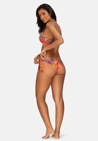 Kobieta w kolorowym, kwiatowym bikini, poprawiająca boczne wiązanie dołu, stojąca boso na tle prostej białej ściany.