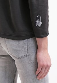 Felpa nera con collo rotondo e maniche lunghe, con un motivo di polpo ricamato bianco sulla manica sinistra. Jeans in denim grigio.