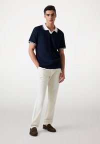 Polo navy con cuello y ribetes de manga blancos, combinado con pantalones crema y mocasines oscuros con flecos. Textura suave, corte casual.