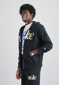 Čierna mikina na zips s kapucňou so zlatým logom "Nike", doplnená o biele tričko s modrým textom a čierne tepláky so zlatým nápisom "nike".