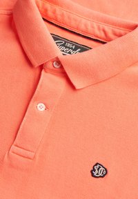 Polo couleur corail orange avec deux boutons blancs, un col plié, et un petit logo brodé foncé sur la poitrine.