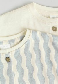 Twee baby T-shirts, één in roomkleur en één met een blauw golfpatroon, met ronde halslijnen en knopen aan de voorkant op een textuurstof.