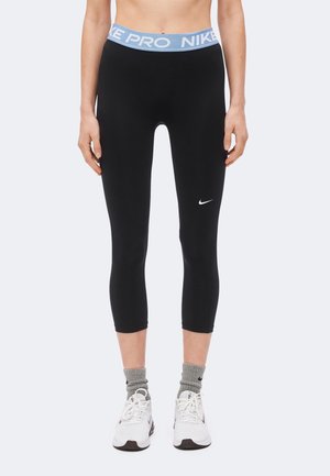 Vrouw draagt zwarte Nike Pro cropped leggings met blauwe tailleband, grijze sokken en witte Nike sneakers, staand tegen een witte achtergrond.