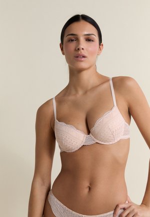 Femme aux cheveux foncés portant un soutien-gorge en dentelle rose clair et des sous-vêtements assortis, debout devant un fond beige uni.
