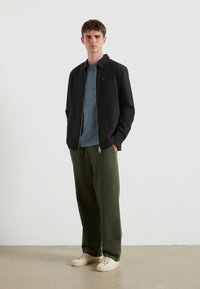 Junger Mann steht mit den Händen in den Taschen, trägt eine schwarze Zip-Jacke, ein blaues T-Shirt, olivgrüne Hosen und beigen Sneakers vor einem einfarbigen Hintergrund.