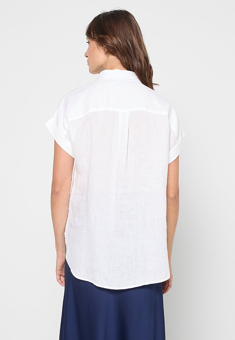 Camicia bianca di lino a maniche corte con colletto, caratterizzata da una leggera texture e un orlo arrotondato, patta con bottoni e dettaglio di cucitura sulla spalla posteriore.