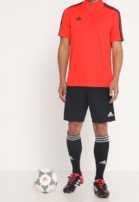 Miesjalkapalloilija, joka pukeutunut punaiseen Adidas-paitaan, mustiin shortseihin, mustiin polven yli ulottuviin sukkiin ja nappulakenkiin, seisoo valkoisen Adidas-jalkapallon vieressä.