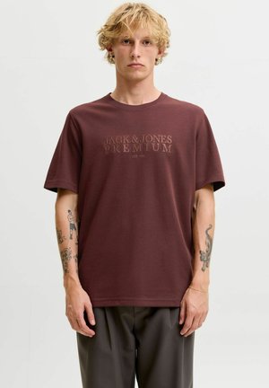 Homme blond avec des tatouages portant un t-shirt marron Jack & Jones Premium et un pantalon gris, debout devant un fond uni.