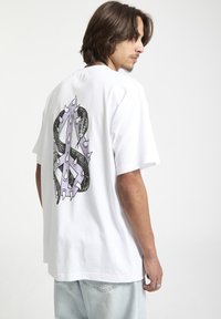 T-shirt en coton blanc avec un design graphique au dos représentant un motif de serpent violet et noir, entouré d'accents en piques.