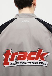 Personne portant une veste grise et noire avec le logo "bareen" près du col et un grand texte rouge "track" avec un slogan dans le dos.