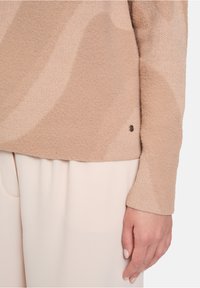 Beige Pullover aus weichem Strick mit wavy Muster und einem subtilen Logo-Design. Schmal zulaufende Ärmel und entspannte Passform, kombiniert mit hellen Hosen.