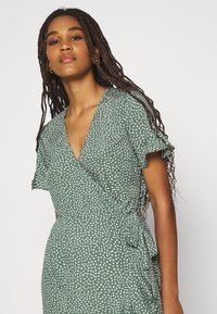 Vestido verde de envoltório com bolinhas brancas, mangas curtas e fluidas, decote em V profundo e detalhe de cintura amarrada. Textura de tecido suave.