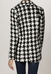 Blazer mit Hahnentrittmuster in Schwarz und Weiß, mit strukturiertem Design, aufgeschlagenem Revers und langen Ärmeln. Die Textur wirkt glatt und gewebt.