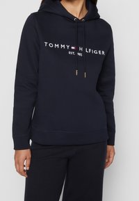 Tmavě modrá mikina s kapucí vyrobená z měkkého materiálu, se předním emblémem s nápisem "TOMMY HILFIGER EST. 1985" v bílé barvě a zlatými akcenty na šňůrkách.