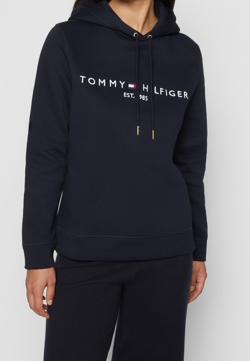 Tmavě modrá mikina s kapucí vyrobená z měkkého materiálu, se předním emblémem s nápisem "TOMMY HILFIGER EST. 1985" v bílé barvě a zlatými akcenty na šňůrkách.