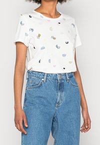 Camiseta blanca de manga corta con patrones abstractos coloridos en diversas formas. Combinada con unos vaqueros azulados de tiro alto. Material de algodón suave.