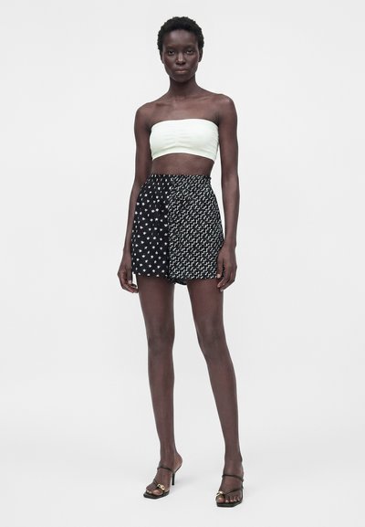 Γυναίκα με σκούρο δέρμα που φοράει λευκό τιραντέ τοπ, μαύρο σορτς με ψηλή μέση με σχέδια αστέρια και κύματα και μαύρα σανδάλια με τακούνι.