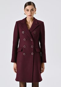 Cappotto bordeaux doppiopetto con grandi bottoni in tonalità argento, colletto a scolla, tasche laterali e silhouette sartoriale. Tessuto dalla texture liscia.