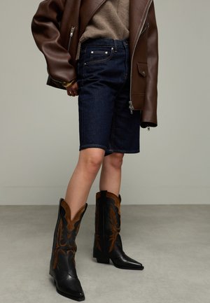 Persoon in een donker denim short, bruine trui, bruin leren jack en zwart-bruin cowboylaarzen die op een grijze vloer staat.