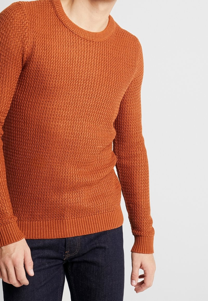 Man i en åtsittande texturerad rust orange stickad tröja med långa ärmar och mörkblå jeans, som står mot en neutral bakgrund.