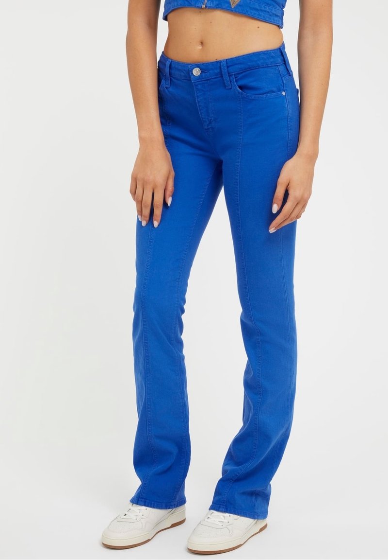 Femme portant un jean droit bleu vif et des baskets blanches, debout devant un fond blanc uni.