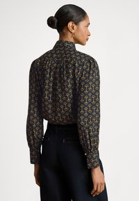 Crna bluza s ponovljenim žutim grafičkim uzorkom, visokim ovratnikom i naboranim rukavima. Kombinirana s tamnim trapericama.